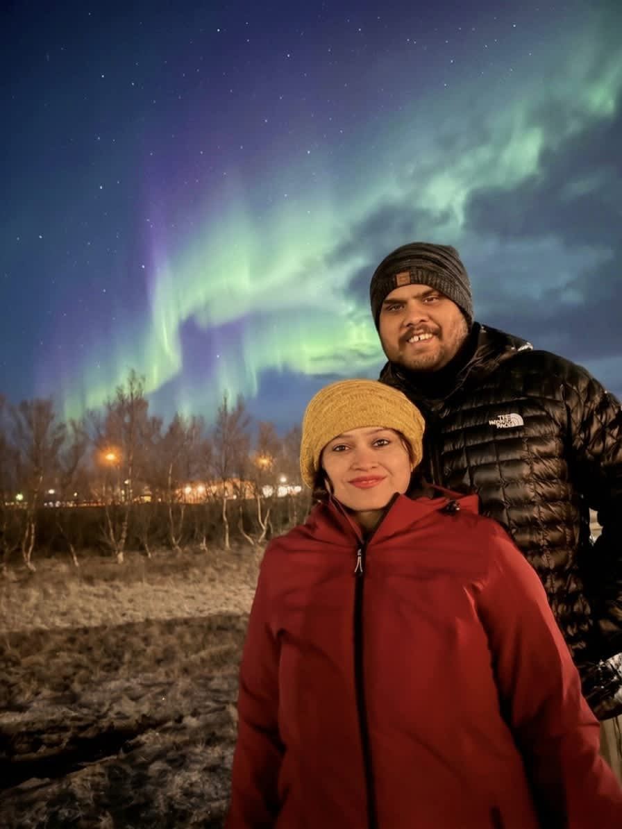 Sucheta and Utsav under the northern lights — green and purple aurora, bare trees, town lights behind