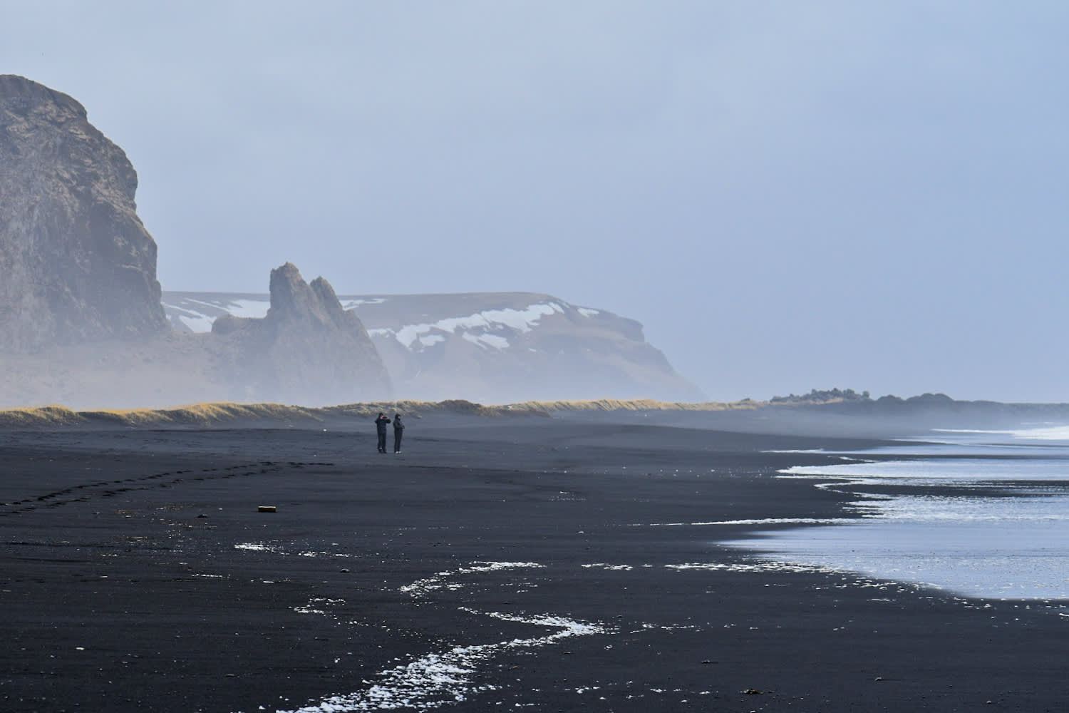 Two tiny figures on the vast misty black beach — the scale of Iceland